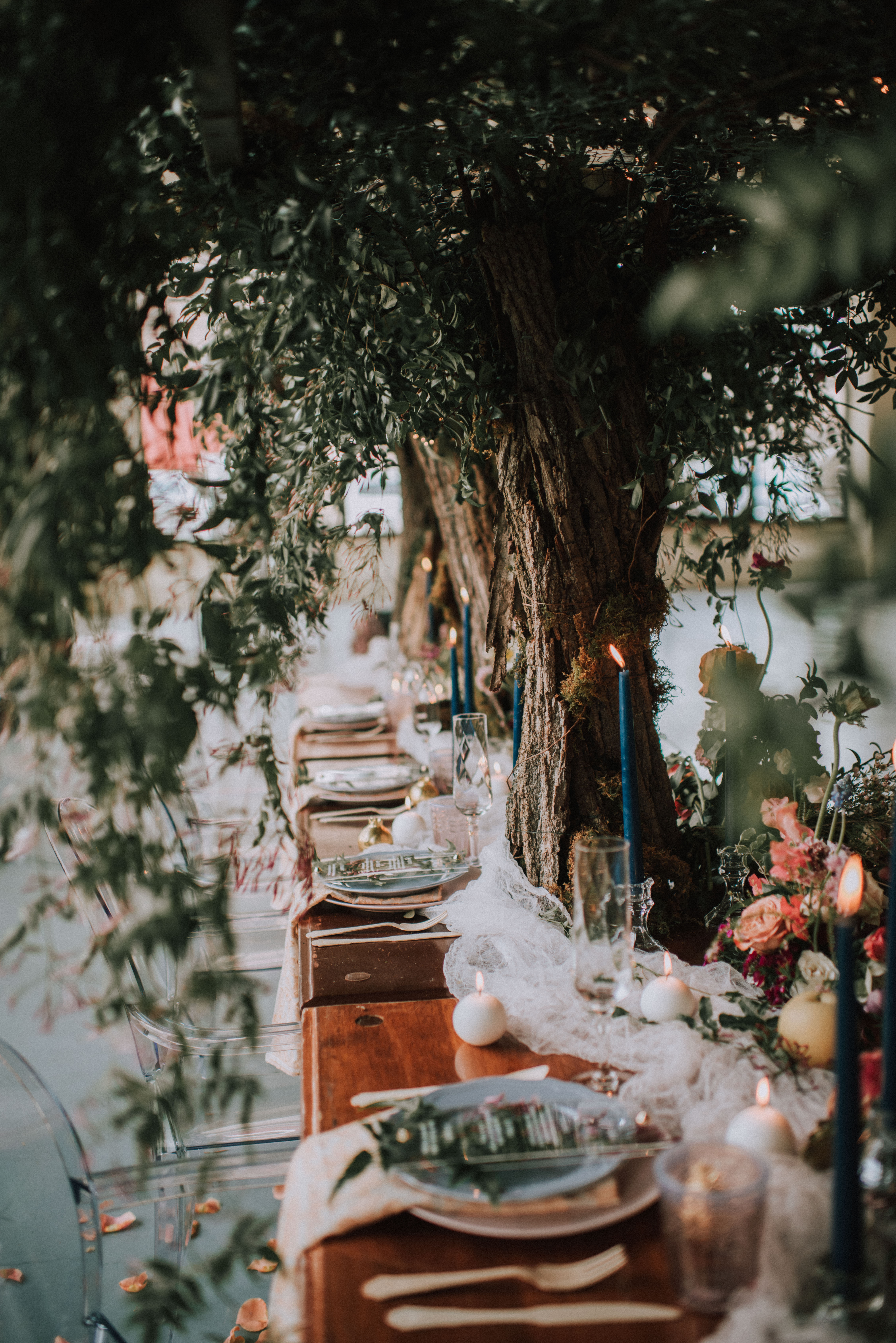 A beautifully set outdoor table beneath lush greenery, adorned with blue candles, elegant dinnerware, and small decorative elements. The setting exudes a rustic and romantic ambiance, perfect for an intimate gathering or event.
